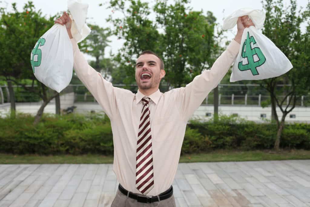 Caucasian man wearing tie holding money bags over his head cheering