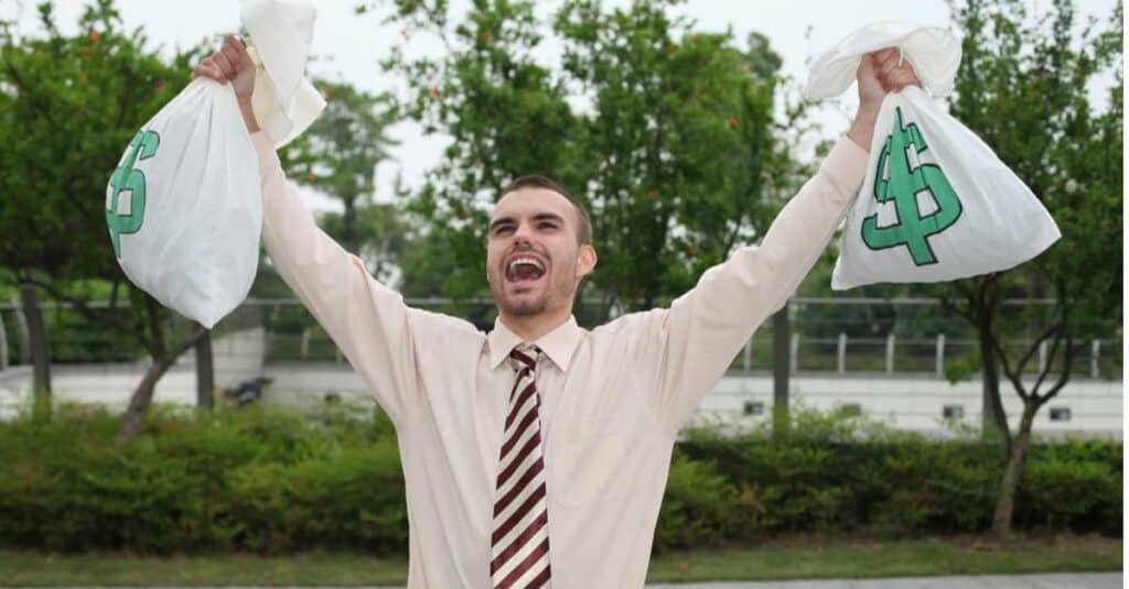Caucasian man wearing tie holding money bags over his head cheering