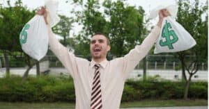 Caucasian man wearing tie holding money bags over his head cheering