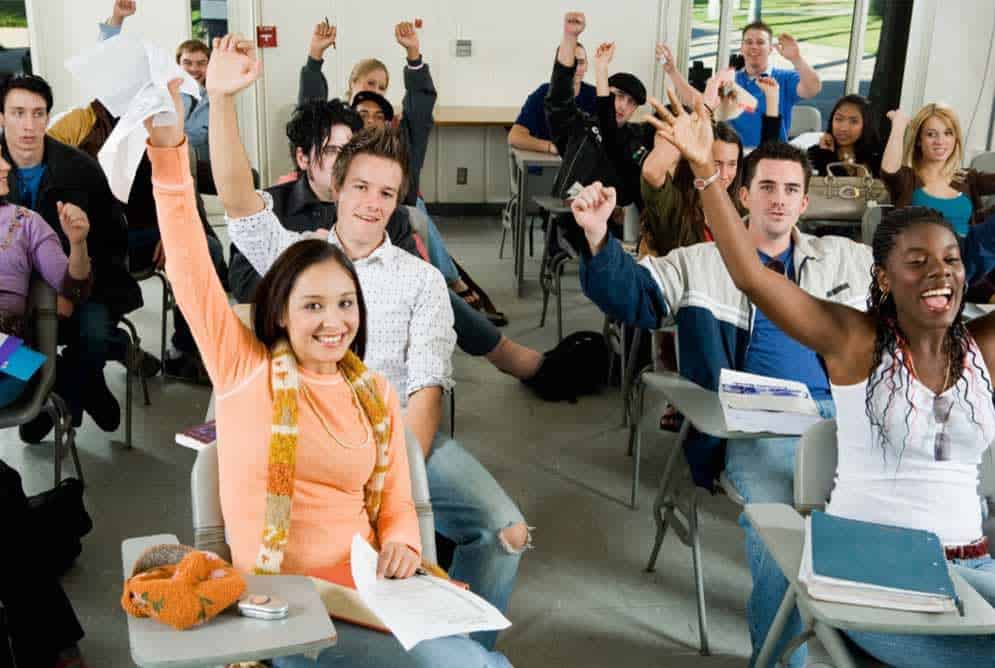 diverse students in classroom sitting in desks with hands up