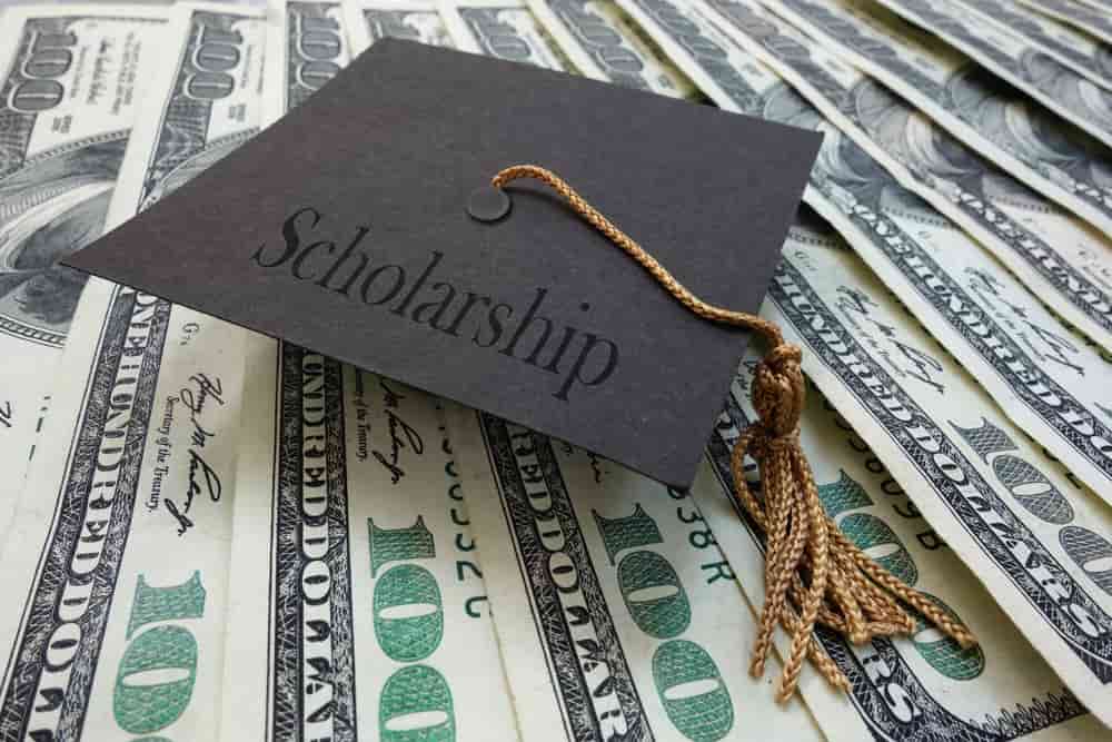 black graduation cap with text scholarship on top of many 100 dollar bills
