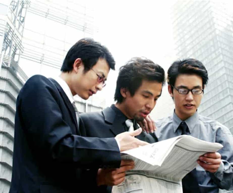 Three asian men looking at newspaper stock market update in city with buildings in background