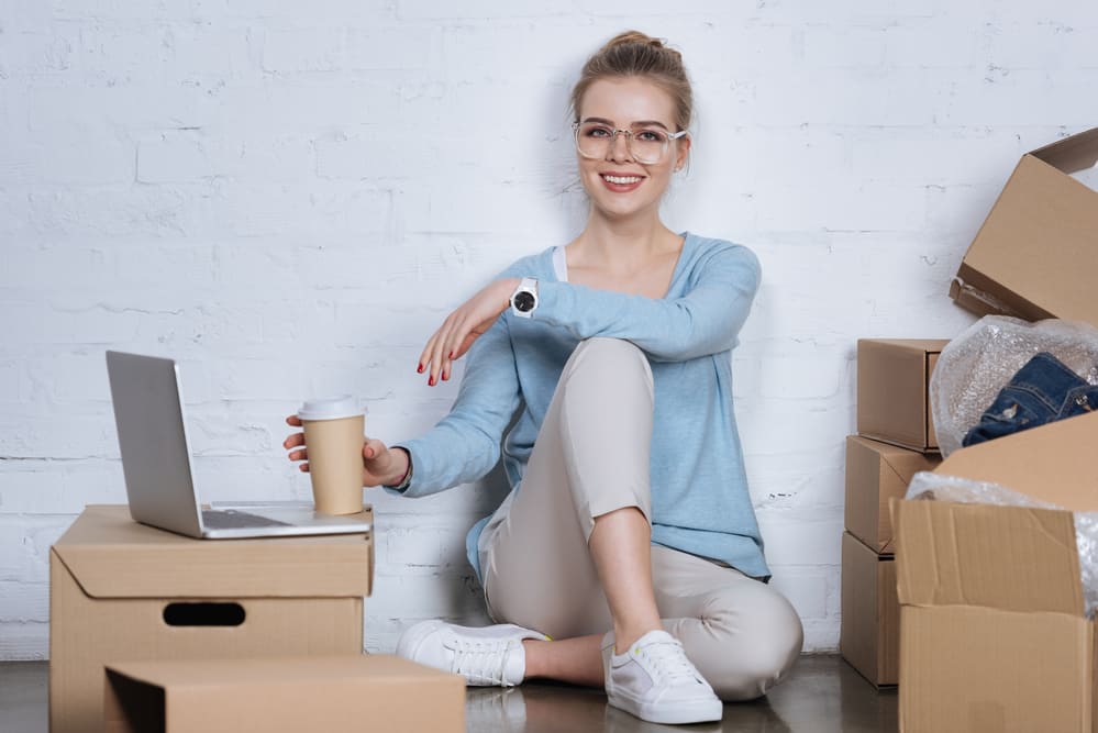 Smiling entrepreneur with coffee to go sitting on the ground with a coffee on unpacked boxes for her online business for teens