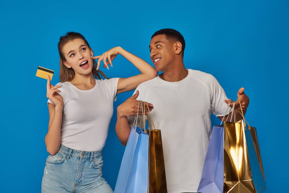 Excited interracial couple holding shopping bags and credit card on blue backdrop