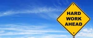 Yellow diamond-shaped sign with black text 'Hard Work Ahead' against blue sky.