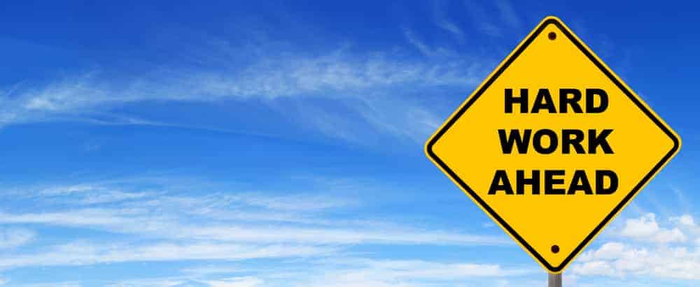 Yellow diamond-shaped sign with black text 'Hard Work Ahead' against blue sky.