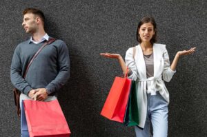 Caucasian girl and caucasian man with red shopping bags leaning on a grey wall. Woman has hands out to side as queston of whats a reasonable clothing budget for a teenager