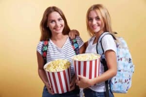 Two smiling teenage girls holding striped popcorn buckets with backpacks.