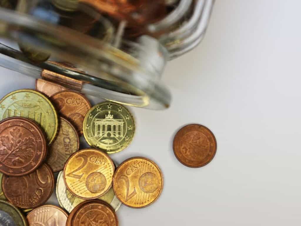Euro coins spilling from a glass jar on a white surface.
