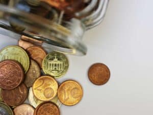 Euro coins spilling from a glass jar on a white surface.