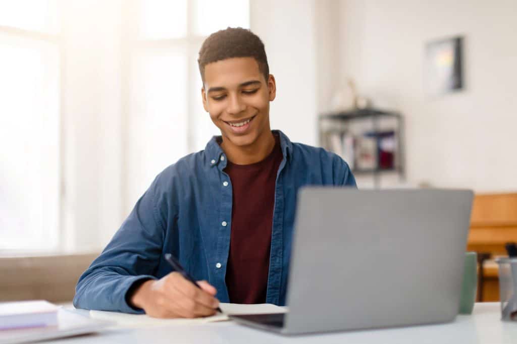 Smiling teenage boy taking notes at his laptop, representing the Gemini Notebook guide for teens