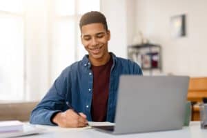 Smiling teenage boy taking notes at his laptop, representing the Gemini Notebook guide for teens