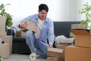 Young man unpacking cardboard moving boxes on the floor of a bright new apartment